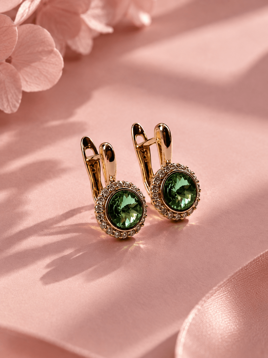 Gold-toned earrings with a green center stone and a frame of clear stones on a pink background