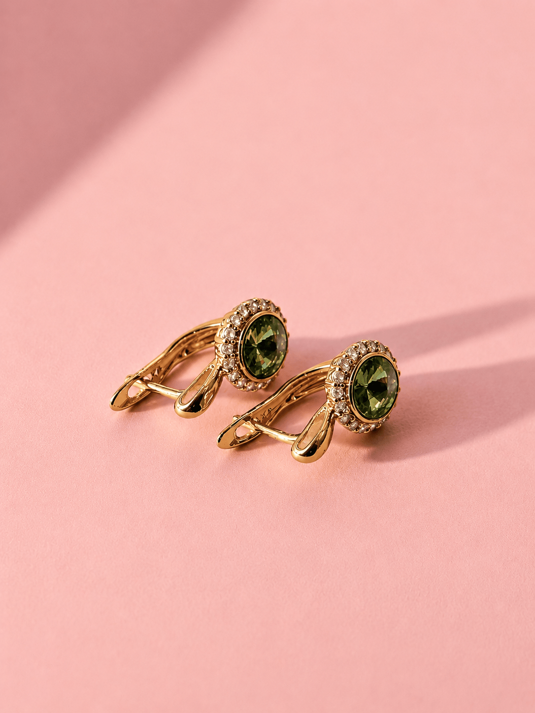 A pair of gold tone earrings with green center stones and English clasp on a pink background